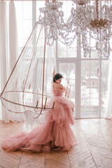 young woman with short black hair in pink princess dress with train is sitting on the crystal chandelier near huge panoramic window background luxury style. fashion concept, free space