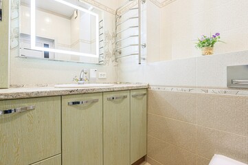 well-lit bathroom features light-green cabinetry, a marble countertop with a sink, and a heated towel rack. A mirror with LED lighting hangs above the sink.