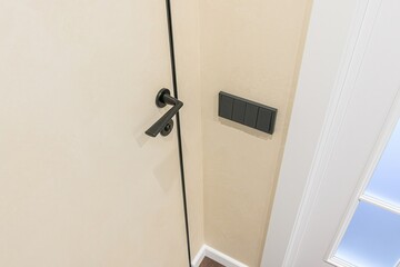 close-up shot of a door with a black handle and frame, next to a wall with a gray light switch panel, adjacent to a white framed window or doorway.