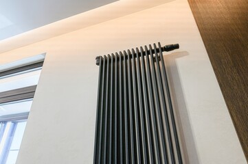 Close-up of a modern black radiator against a light wall, adjacent to a window and dark wood panel