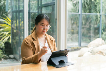 Asian Businesswoman A woman sits at a table, using a tablet in a serene, green environment filled...