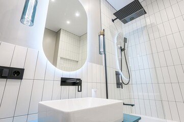 Modern bathroom with white tiles, black fixtures, and an oval mirror with integrated lighting....
