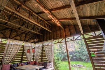 Rustic wooden gazebo with fabric curtains, colorful pillows, and string lights. View of trees outside