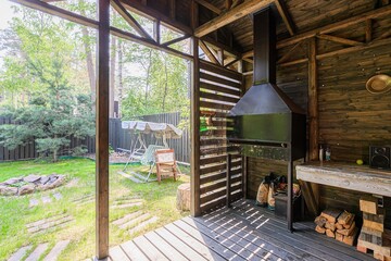 rustic outdoor space features a wooden structure with a grill, wood storage, and a view to a garden with a swing and trees.
