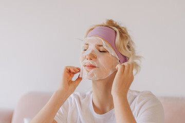 Young teenager touching face with moisturizing sheet mask, posing in room at home