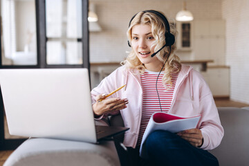Smiling blonde young teenager studying online with headset and laptop sitting at home
