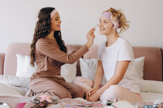 Happy pretty young teenager applying makeup for her friend sitting in bedroom at home