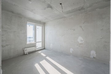 Bare, light-filled room in renovation with exposed concrete ceiling, plastered walls, and smooth...