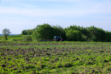 A blue tractor with a trailer stands in a field among green grass and prepared soil. Trees and a light sky are visible in the background.