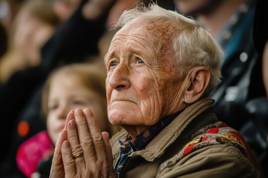 Elderly man with clasped hands looks upward in hope, faith, or concern in thoughtful reflection, surrounded by others at an event