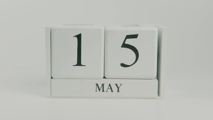 A hand flips a wooden calendar from May 15th to May 16th on a white background, signaling another day in the month