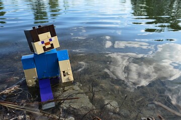 Naklejka premium LEGO Minecraft Steve walking out of shallow water of large spring fish pond, remains of aquatic plants under his feet and reflection of clouds visible. 