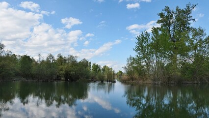Panoramic landscape with larger fish pond surrounded by broadleaf trees, reflection of blue skies with clouds visible on calm water surface. Location western Slovakia, central Europe. 