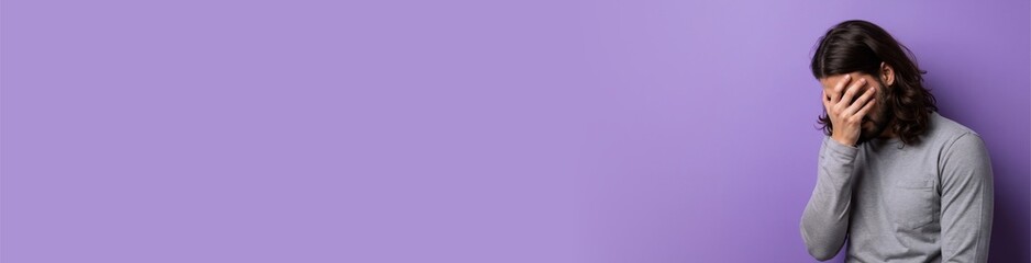 man with long hair leaning on violet wall, hand over face, frustrated body language