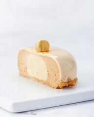 Cut view of individual praline hazelnut mousse cake with glossy glaze and crunchy base. Smooth interior layers and golden topping. Served on white plate over marble background. 
