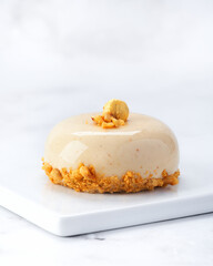 Elegant individual praline hazelnut mousse cake with glossy glaze, golden topping and crunchy nut base. Side view on white plate over marble background with copy space. Perfect for packaging or menus.