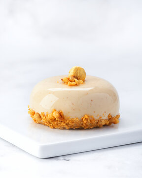 Elegant individual praline hazelnut mousse cake with glossy glaze, golden topping and crunchy nut base. Side view on white plate over marble background with copy space. Perfect for packaging or menus.