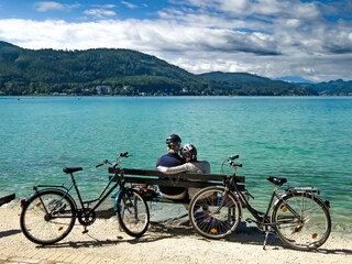 Sporty Senior Couple With Bicycles Sit On Bench At Lake Woerthersee In Carinthia, Austria