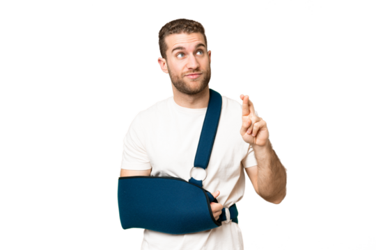 Young handsome blonde man with broken arm and wearing a sling over isolated chroma key background with fingers crossing and wishing the best