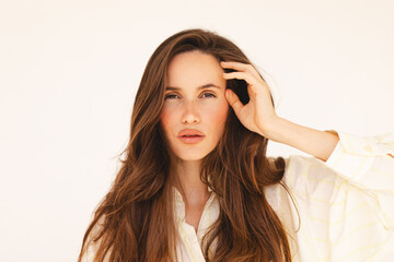 Fototapeta premium Brunette woman with long hair, wearing a white shirt, looking intensely at the camera with a hand near her face. Perfect for beauty, elegance, self-reflection, identity, emotion, or fashion concepts.