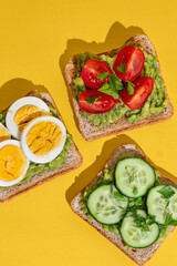 Delicious healthy bread toasts with fresh avocado, boiled eggs, cherry tomato and cucumber placed on bright yellow background