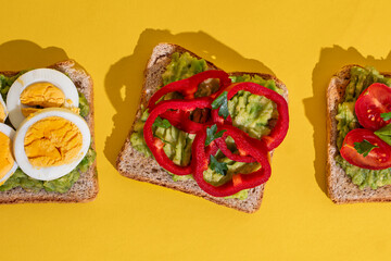 Delicious healthy bread toasts with fresh avocado, boiled eggs, cherry tomato and cucumber placed on bright yellow background