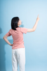 Young Asian woman standing against a blue wall and pointing upwards with her right hand in a...