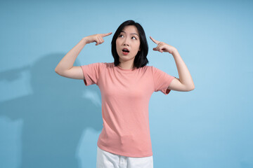 Young Asian woman expresses curiosity and surprise while pointing to her head against a light blue...