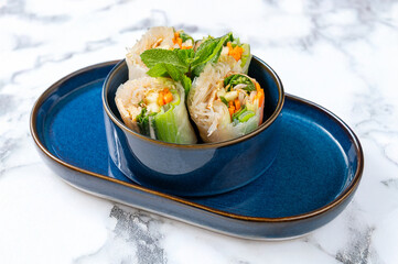 Vietnamese rolls with rice leaf, soy noodles, vegetables and chicken, served in blue ceramic bowls on a white veined marble background. Cool, minimalist photo, ideal for Asian cuisine themes.