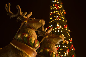 Christmas reindeer figurines adorned with festive harnesses, set against backdrop of tall, illuminated Christmas tree.