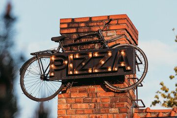 A retro-style pizza sign made of marquee lights is mounted on a brick wall, creatively integrated with an old bicycle.