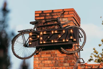 Selbstklebende Fototapeten A retro-style pizza sign made of marquee lights is mounted on a brick wall, creatively integrated with an old bicycle. © Emvats