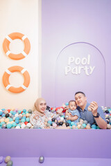 Happy young family playing in a colorful ball pool during a fun indoor "Pool Party" photo session in studio. Parents and baby are smiling and enjoying time together. 