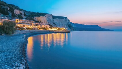 Coastal town at twilight.  Tranquil beach scene at dusk with illuminated buildings reflected in calm water.  White cliffs line the horizon
