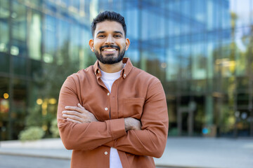 A smiling man stands confidently with arms crossed, posing in front of a modern glass building. The outdoor setting captures a professional and approachable vibe.