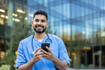 A smiling healthcare professional in scrubs uses a smartphone outside a modern building. His...