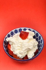 A blue and white patterned bowl holds fresh strawberries generously covered with white whipped cream against a vibrant red background