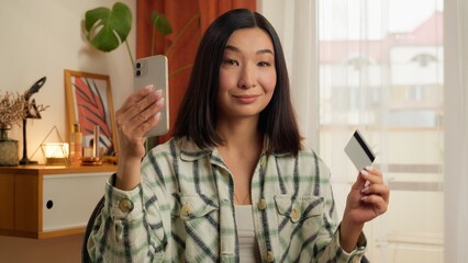 Happy Asian Korean woman holding credit card mobile phone smartphone female smiling businesswoman home indoors online payment shopping discounts client customer lady app Chinese Japanese girl business
