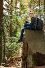 Fototapeta premium A boy sits on a large rock in the forest during a hike, wearing casual clothes and holding a backpack. Sunlight filters through the trees, creating a peaceful outdoor atmosphere. 