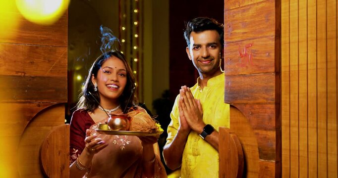 Praying together with puja thali, Indian young couple in traditional ethnic attire doing namaskar at home entrance for Hindu Diwali festival or welcoming ceremony while entering new house joyfully