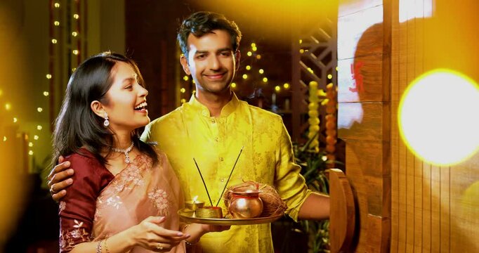 Drawing swastik showing house key with puja thali, Indian young couple in traditional wear greeting while welcoming camera into their new home on Hindu Diwali festival joyfully