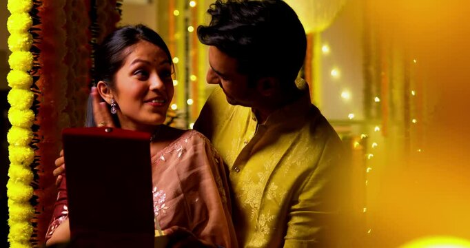 Indian husband gifts necklace to wife as surprise on Diwali festival night while standing together joyfully against decorated marigold garland wall sharing love laughter with playful festive emotions