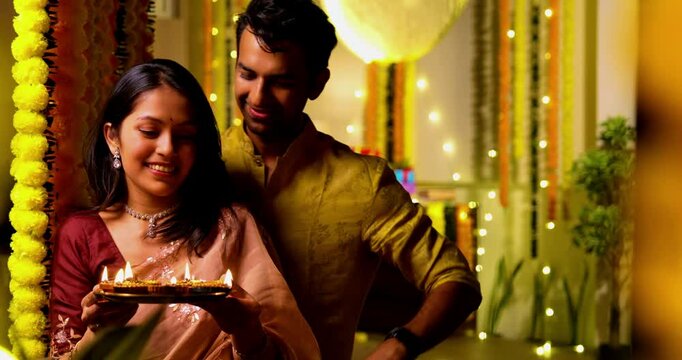 Holding diya showing house key, Indian young couple in festive ethnic wear celebrating first Diwali in new home, smiling standing in front of colorful garlands with joy togetherness on festival night