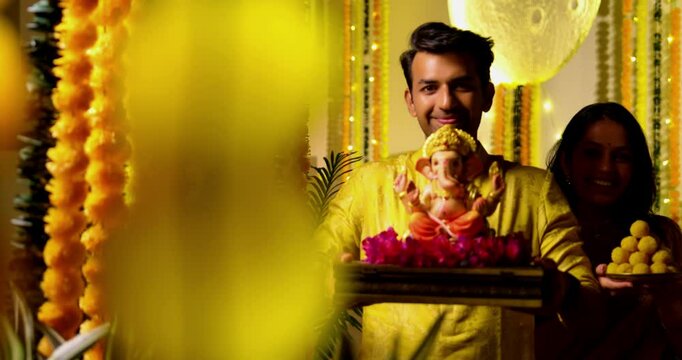 Indian Couple Welcoming Ganesh Idol with Joy, Young husband wife in Traditional Attire Holding Ganpati Bappa Idol, laddu thali While Smiling Chanting Bappa Morya During Ganesh Festival Celebration