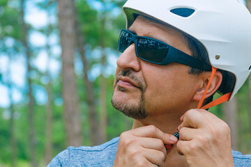 Preparation to Cycling: A portrait of a sport enthusiast fastening strap of a bicycle helmet against the background of an urban park. Photo is part of a series that highlights theme of healthy living.