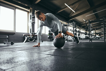 Horizontal photo of strong guy in sportswear doing push up exercise with one hand on medicine ball during intense fitness workout in modern gym