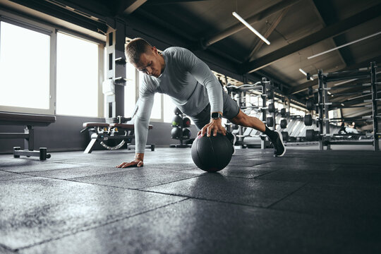 Horizontal photo of strong guy in sportswear doing push up exercise with one hand on medicine ball during intense fitness workout in modern gym