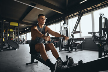 Handsome male practicing on rowing machine in modern gym. Athletic young man working out on rowing machine at fitness center. Weight reduction and cardio workout concept