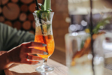 Colorful summer cocktail drink. Orange beverage with mint and ice. Hand holding refreshing tropical drink. Wooden table and warm sunlight. Refreshing bar scene with exotic cool refreshment.