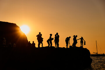 Fototapeta premium Silhouettes of people at sunset on a mountain or cliff, enjoying the view and relaxing in a warm golden light. Summer social moment in nature.